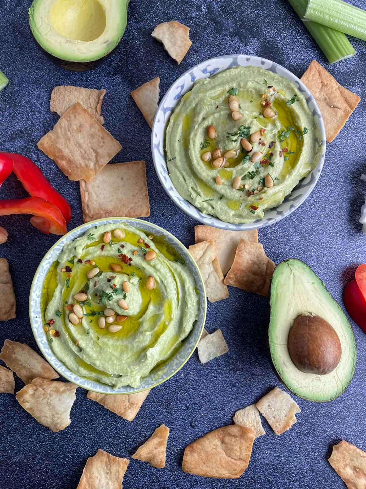 Two bowls of creamy avocado hummus surrounded by pita chips and veggies.